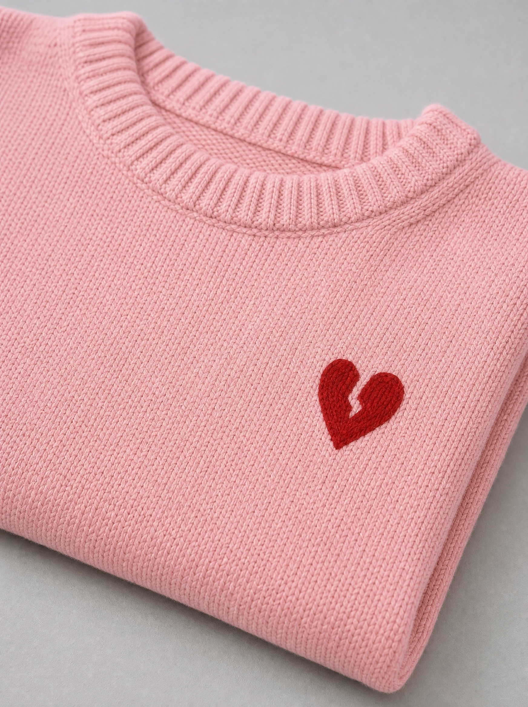 Close up of the ribbed crew neck and the small red heart motif on a pink knit sweater