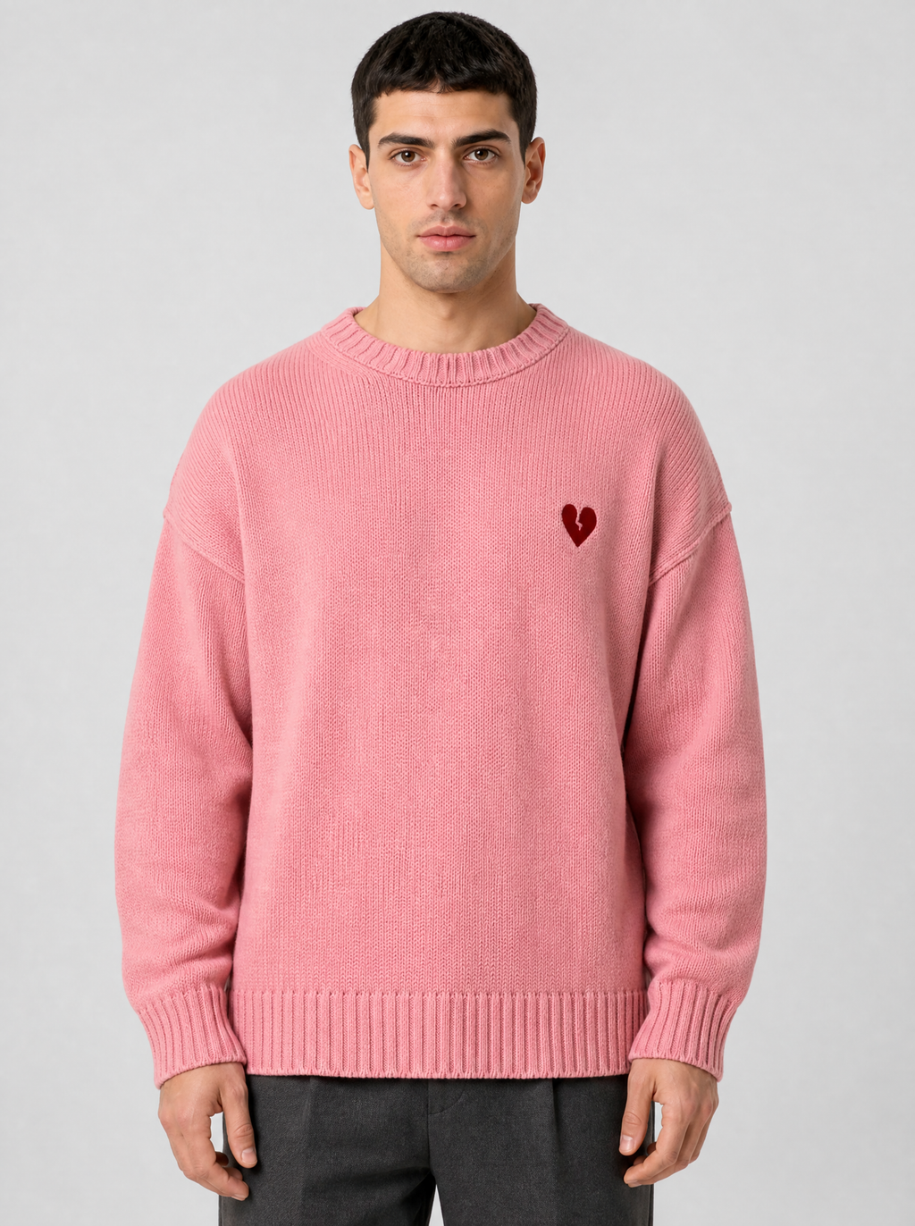 Person wearing a pink knit sweater with a small red heart at the chest, front view on a light grey studio background
